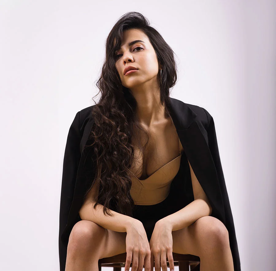 A woman with long, wavy hair, sitting confidently on a chair, wearing a black blazer draped over her shoulders and a nude top, against a white background - Botox in Chicago, IL