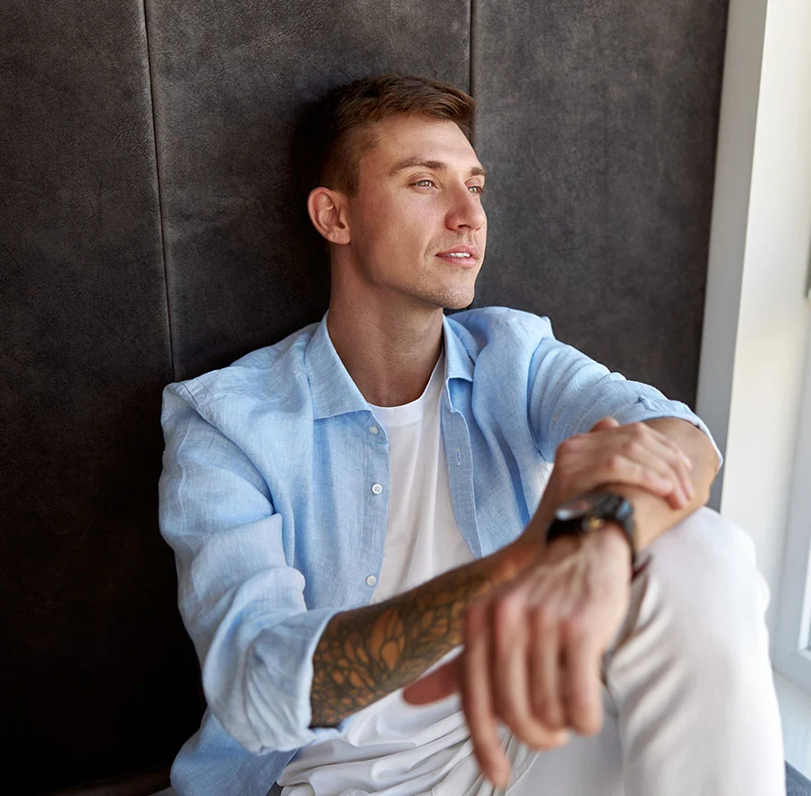 A blonde man looking out the window, against a grey textured wall, wearing a white button up, with tattoos - Earlobe Repair Procedure in Chicago, IL