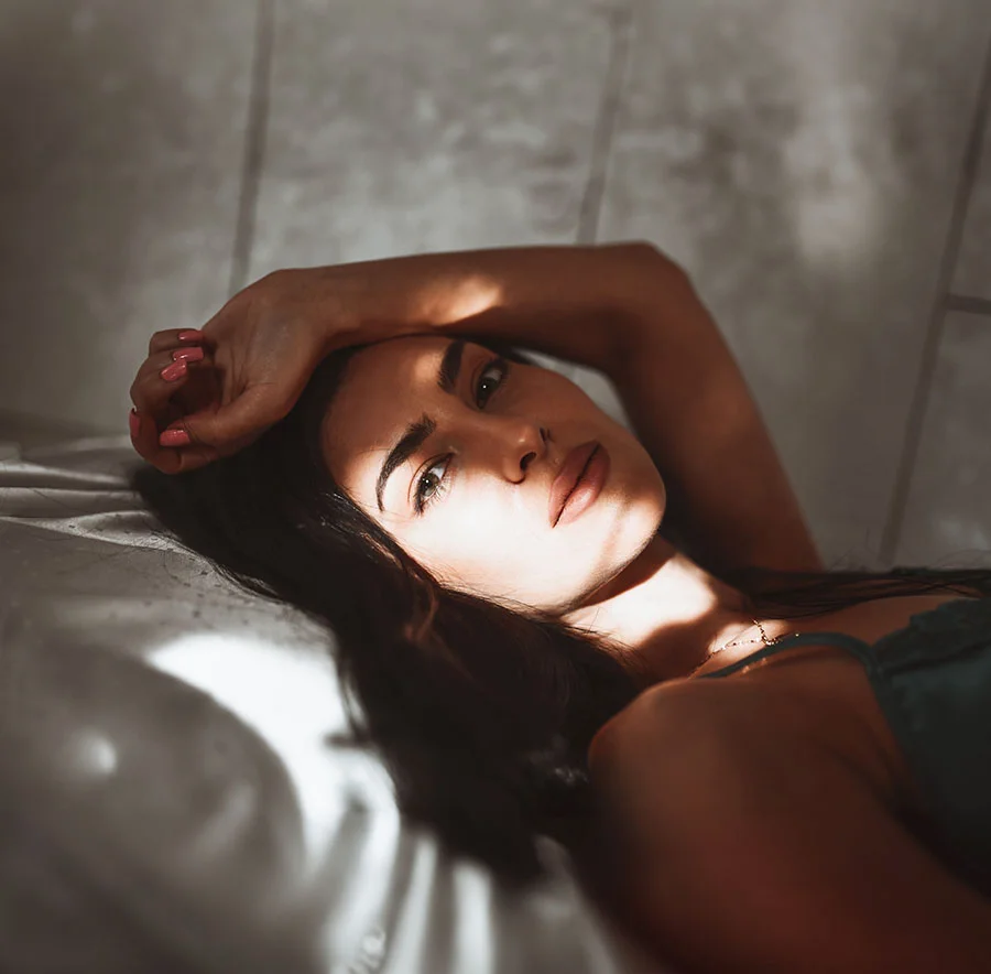 A woman lying in bed, with short brown hair, as the sun shines on her in a dimly lit room, her arm raised over her head - Eyelid Surgery in Chicago, IL