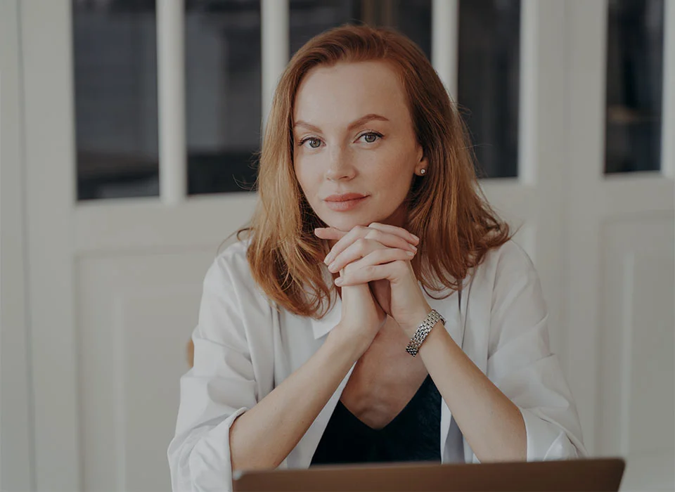 A red haired woman, wearing a white button up shirt over a black tank top, hands rested under her chin - Eyelid Surgery in Chicago, IL