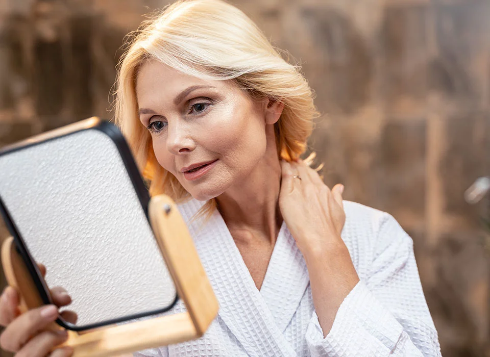 A mature woman with blonde hair examines her reflection in a handheld mirror, wearing a white bathrobe, set against a neutral, softly lit background - Facelift Procedure in Chicago, IL