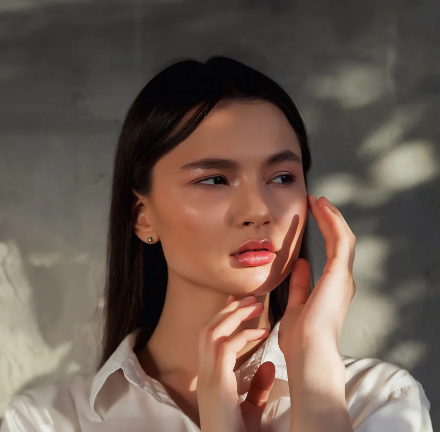 A woman with long dark hair, standing against a textured wall, her face softly illuminated by sunlight, wearing a white blouse, her hands gently touching her face - Functional Rhinoplasty Procedure in Chicago, IL