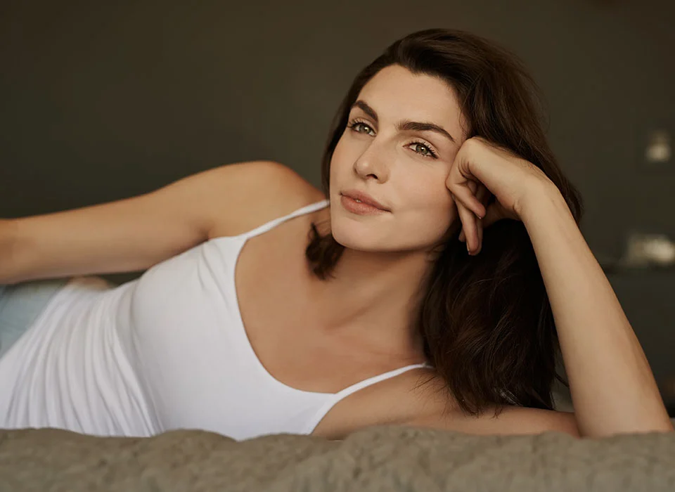 A woman with dark hair, lying on a bed, wearing a white tank top, her head propped on her hand, looking relaxed - Functional Rhinoplasty in Chicago, IL