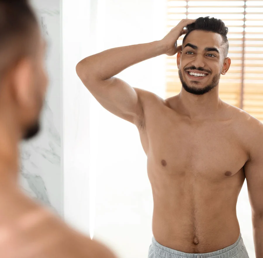 A shirtless man with dark hair and a beard, standing in front of a mirror, smiling confidently while running his hand through his hair - Gynecomastia Procedure in Chicago, IL