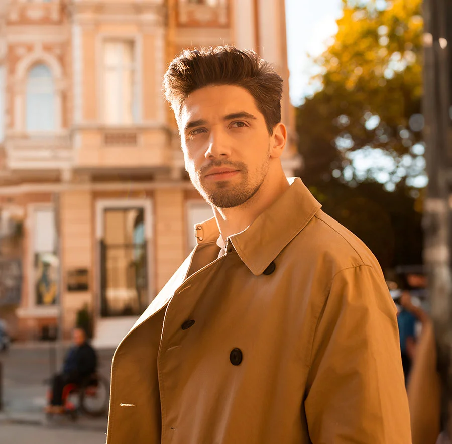 A man with dark hair, wearing a beige trench coat, standing outdoors in a sunlit urban setting - Hair Restoration Procedure in Chicago, IL