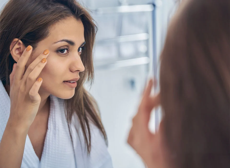 A woman in a white robe, looking at herself in the bathroom mirror, gently touching her face with a focused expression - Surgical Skin Tightening Procedure in Chicago, IL